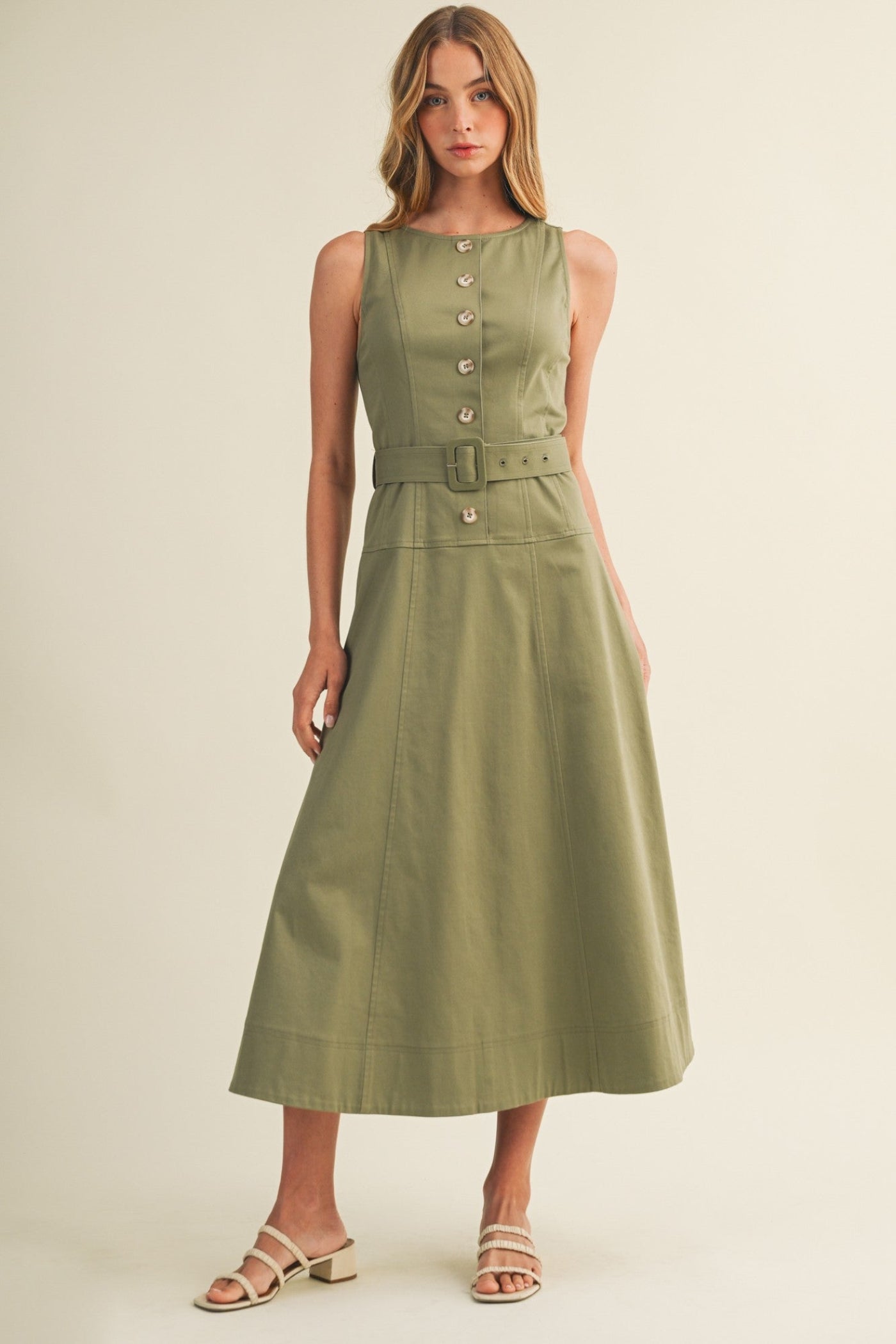 A woman modeling a sleeveless olive green midi dress with a button-front bodice, matching belt, and flared skirt, paired with beige strappy sandals against a plain beige studio background.