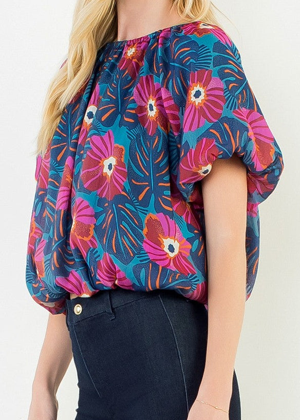A woman modeling a short-sleeve blouse with a vibrant floral and leaf pattern in pink, teal, and orange, paired with dark navy pants, against a plain white studio background.