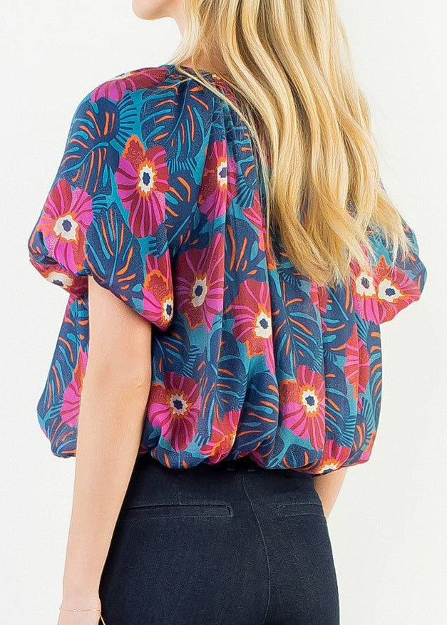 A woman shown from the back wearing a short-sleeve blouse with a colorful floral print and dark blue jeans, photographed against a plain white studio background.