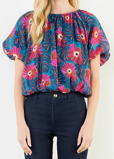 A woman stands against a plain white wall wearing a colorful short-sleeve blouse with a pink, blue, and green floral pattern, paired with dark navy jeans and white heels.