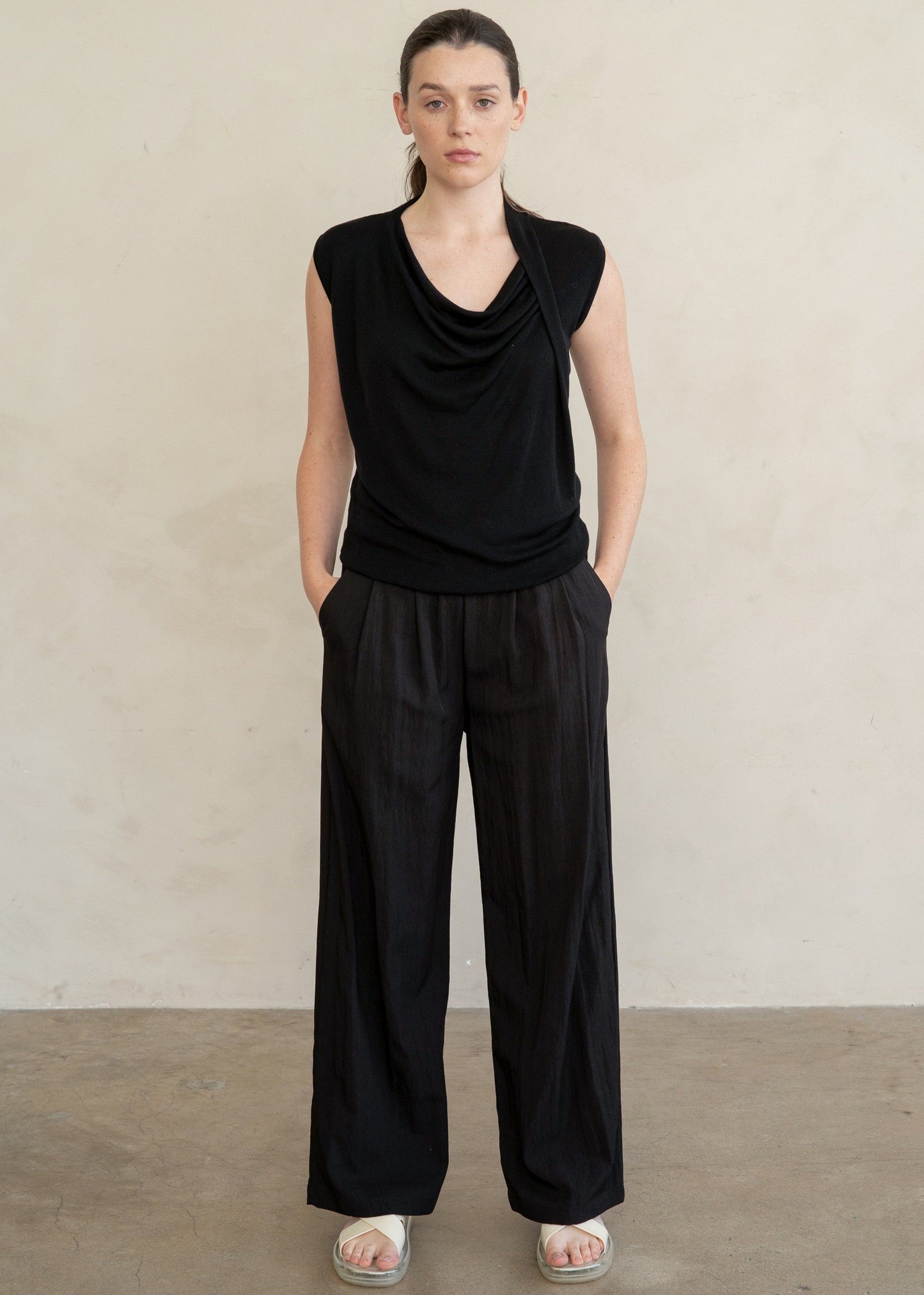 A model stands against a neutral beige wall wearing a black sleeveless cowl-neck top and matching black wide-leg trousers, paired with white slide sandals.